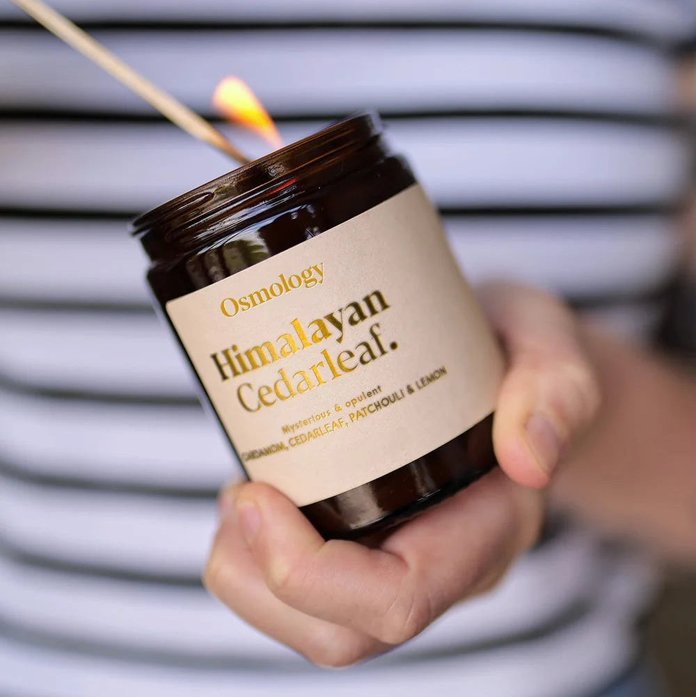 Person holding a lit candle labeled 'Osmology Himalayan Cedarleaf' against a striped background