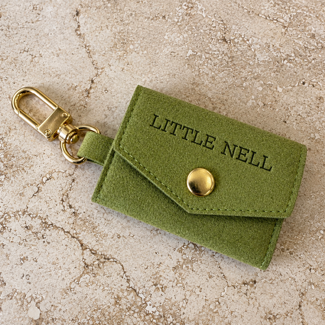 An olive green suede-like key case with a gold-tone clasp and a gold snap button, featuring the text 'LITTLE NELL' debossed on the flap, set against a textured beige stone background.