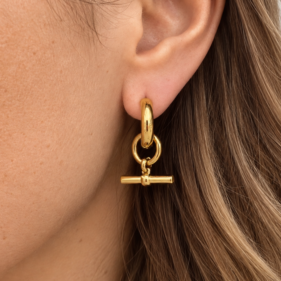 A close-up of a person's ear wearing a polished gold huggie hoop earring with a dangling toggle bar charm.