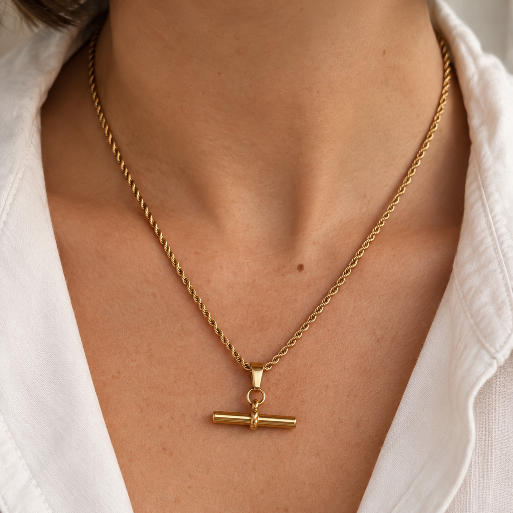 A close-up of a person wearing a delicate gold necklace with a toggle clasp pendant, styled with a white collared shirt.