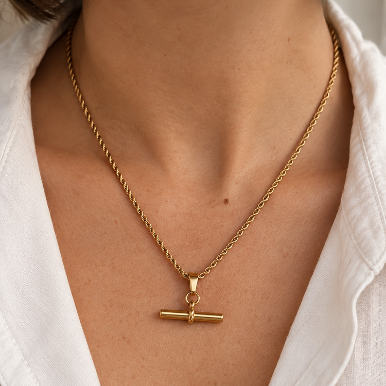 A close-up of a person wearing a delicate gold necklace with a toggle clasp pendant, styled with a white collared shirt.