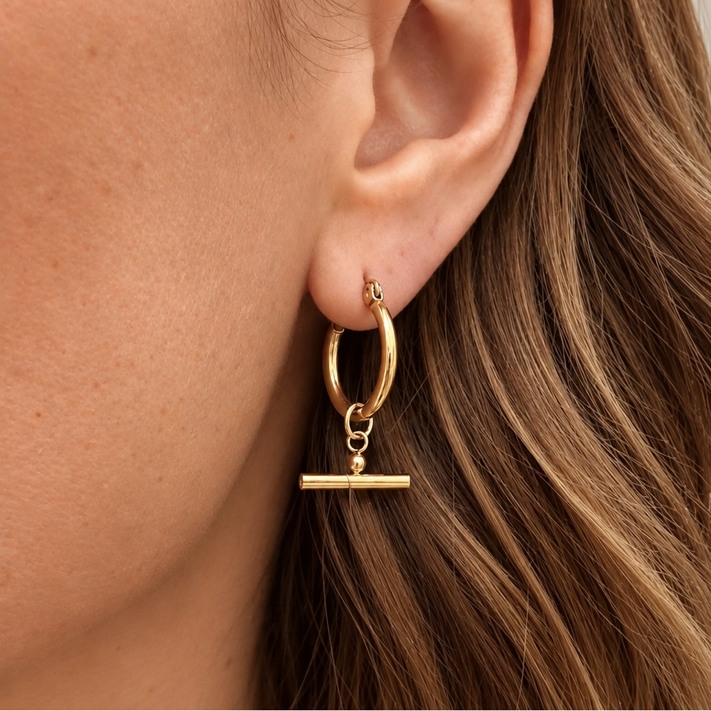 A close-up of a model's ear wearing a gold hoop earring with a dangling toggle bar earring.