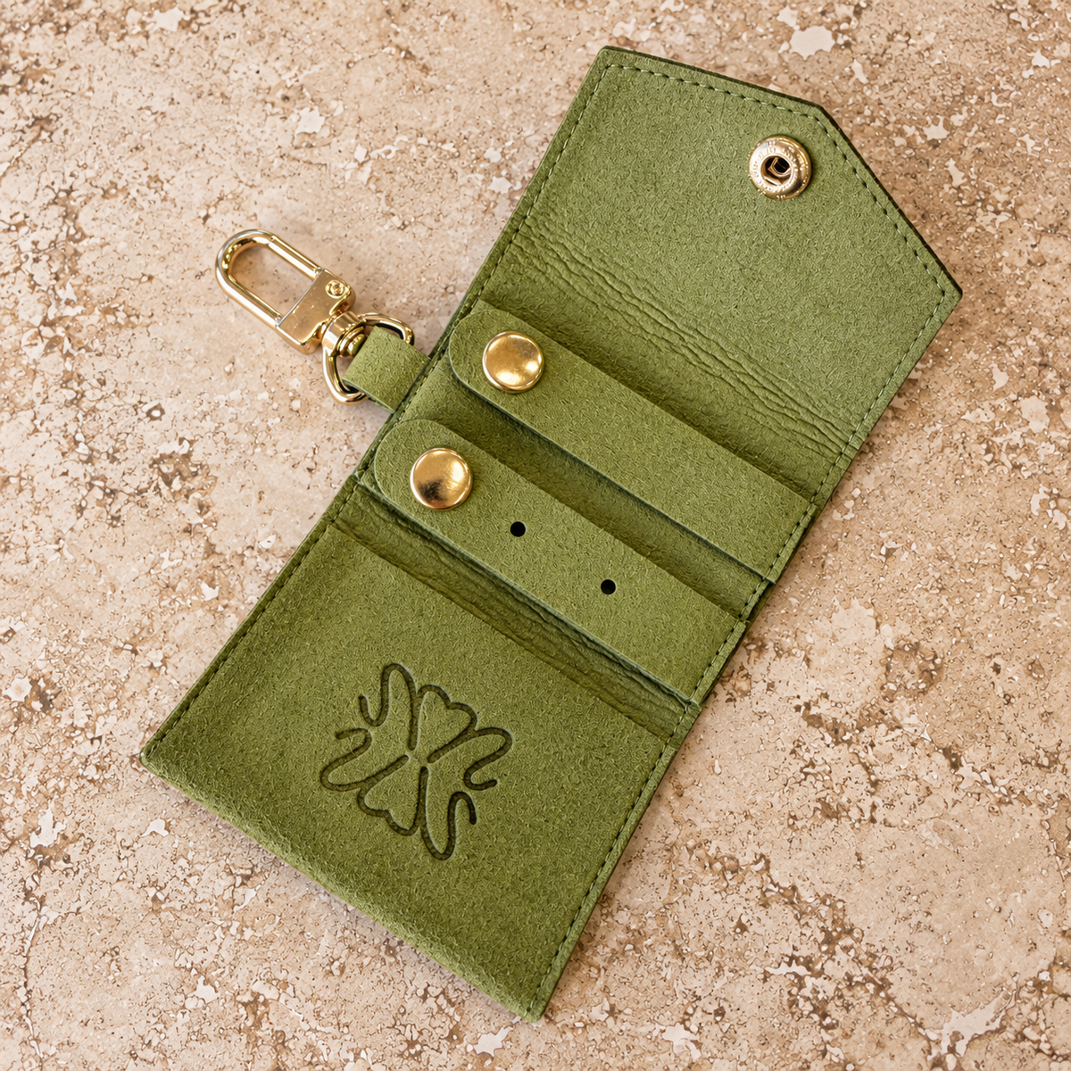 An olive green faux-leather key card holder wallet with a gold-tone snap button, card slots, and a gold clasp, shown on a textured beige stone background.