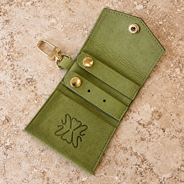 An olive green faux-leather key card holder wallet with a gold-tone snap button, card slots, and a gold clasp, shown on a textured beige stone background.