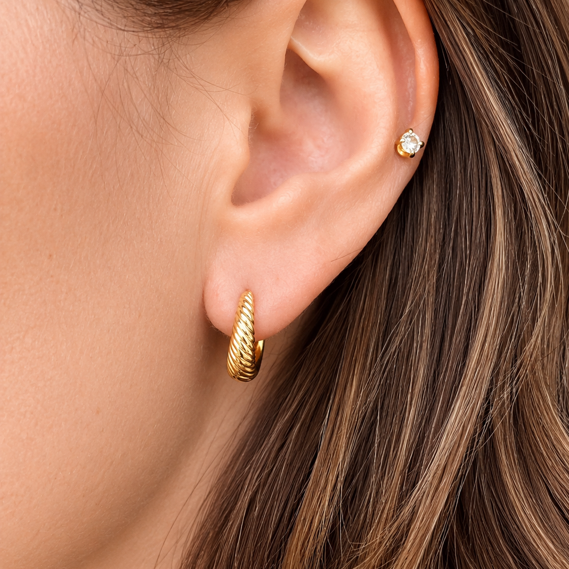 A close-up of a model's ear showcasing two gold earrings: a textured gold huggie hoop on the lobe and a small gold stud with a clear gemstone on the helix.